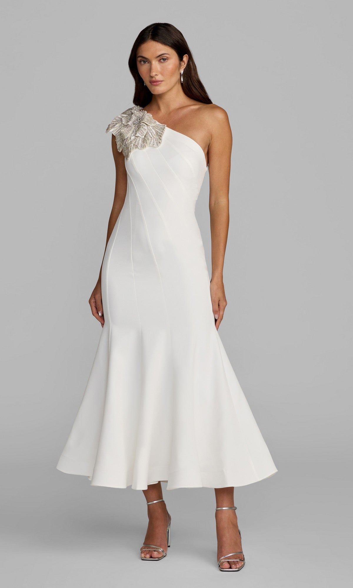 Woman wearing the Mac Duggal 13221 Tea-Length Party Dress in ivory, featuring a crepe one-shoulder design and embellished floral shoulder applique, poses against a plain gray background.